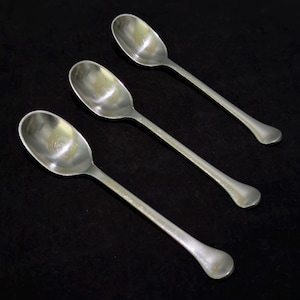 May include: Three silver spoons with oval bowls and long, slender handles are arranged diagonally against a dark background. The spoons have a classic design, reflecting light and creating highlights on their surfaces.