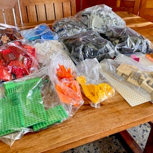 May include: A collection of colorful plastic building blocks in various colors, including red, green, yellow, and blue, are contained in clear plastic bags. The blocks are arranged on a wooden table, ready for construction.