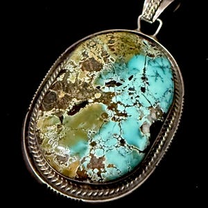 May include: Oval turquoise pendant with a silver setting. The turquoise stone features a mix of blue, green, and brown hues with intricate veining. The pendant has a decorative silver bail and a rope-like border.