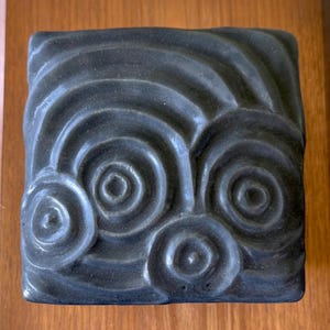 May include: A square, dark gray decorative tile with a raised spiral design. The tile is made of a textured material and is mounted on a wooden surface. The spiral pattern is repeated three times across the tile.