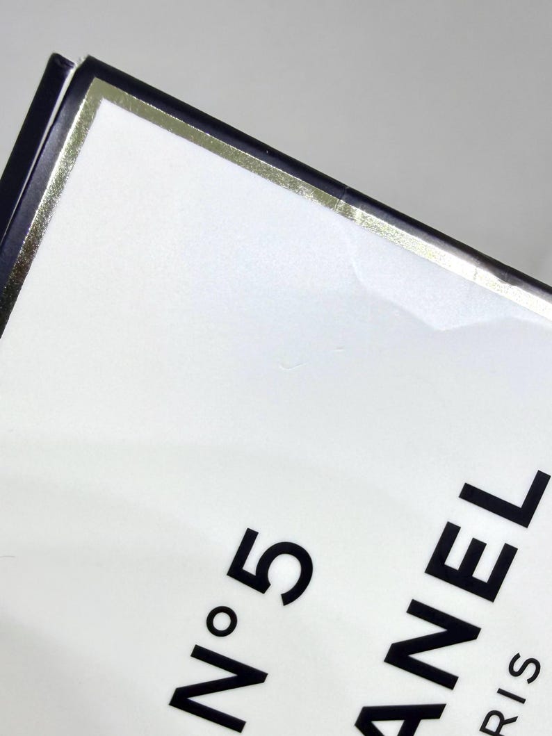 May include: A white rectangular box with a black border and a thin gold metallic trim. The text N&deg;5 is printed in black, with the letters ANEL and RIS partially visible below it, indicating the branding on this fragrance packaging.