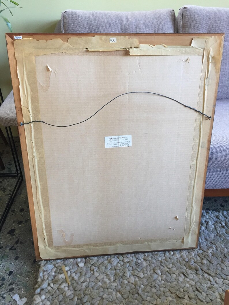 May include: The back of a framed picture with a brown wooden frame and a cardboard backing. The frame has a label that says "44" and a sticker that says "Certificate of Guarantee".