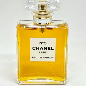 May include: A rectangular glass perfume bottle filled with amber-colored liquid. The white label features the black text N&deg;5 CHANEL PARIS EAU DE PARFUM. The bottle is topped with a clear, faceted, square-shaped glass stopper and a gold-toned collar.