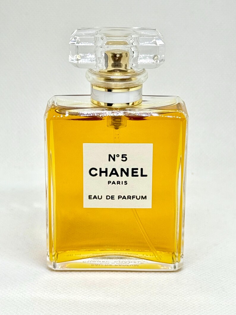 May include: A rectangular glass perfume bottle filled with amber-colored liquid. The white label features the black text N&deg;5 CHANEL PARIS EAU DE PARFUM. The bottle is topped with a clear, faceted, square-shaped glass stopper and a gold-toned collar.
