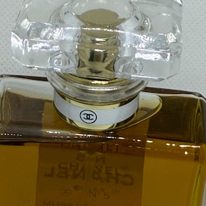 May include: A rectangular glass perfume bottle filled with amber liquid, featuring a clear faceted stopper and a gold-toned neck collar. A white band with a black Chanel logo encircles the neck. The label reads CHANEL N&deg;5 EAU DE PARFUM.