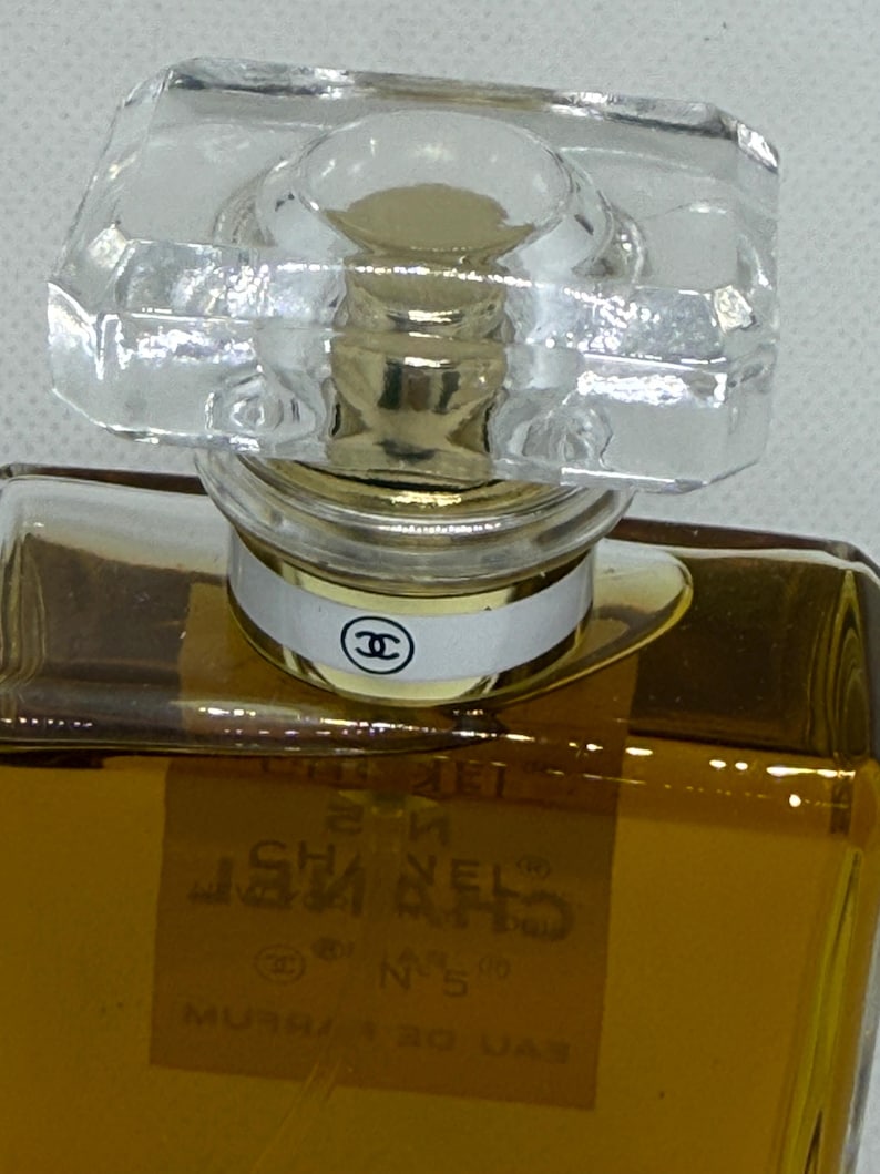 May include: A rectangular glass perfume bottle filled with amber liquid, featuring a clear faceted stopper and a gold-toned neck collar. A white band with a black Chanel logo encircles the neck. The label reads CHANEL N&deg;5 EAU DE PARFUM.