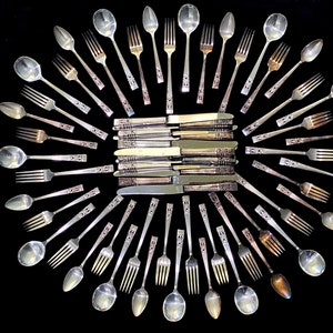May include: A set of silverware arranged in a circular pattern on a black background. The set includes spoons, forks, and knives, all with ornate designs.