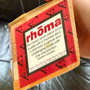 May include: A wooden Soma cube puzzle with a red and white label. The label reads "rhōma" and describes the puzzle as a classic seven-piece puzzle with a rhombic slant. The box is held by a person.