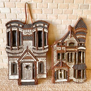 May include: Two ceramic house-shaped wall hangings. The houses are detailed with brown and white paint, featuring bay windows, doors, and architectural details. One has a leather hanging loop.