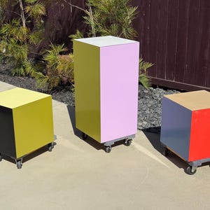 May include: Three colorful, geometric display stands on wheels. One is a tall, rectangular shape with pink and yellow sides. The other two are cube-shaped, with yellow and black, and red, gray, and blue sides. The stands are outdoors.