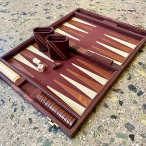 May include: A brown backgammon board set featuring a felt playing surface with alternating cream and brown triangular points. The set includes two brown dice cups, a doubling cube, two white dice, and two sets of circular checkers in cream and brown.