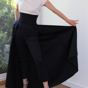 May include: A woman wearing a white blouse with a black belt and a long black skirt. The skirt is a wrap-around style and has a slit in the front. The woman is wearing black heels.