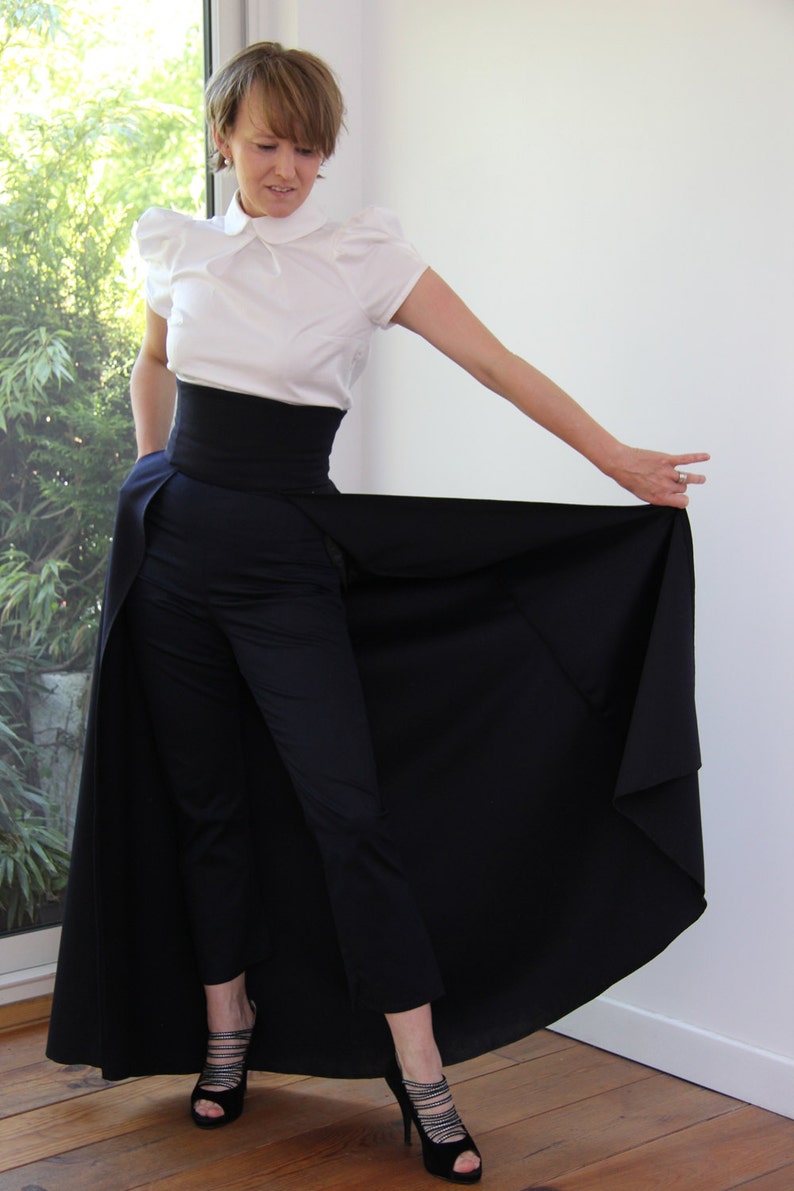 May include: A woman wearing a white blouse with a black belt and a long black skirt. The skirt is a wrap-around style and has a slit in the front. The woman is wearing black heels.