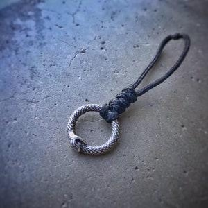 May include: A silver-colored Ouroboros pendant with a snake biting its tail, attached to a black rope. The snake's body has a scale pattern. The rope is knotted at the top and forms a loop.