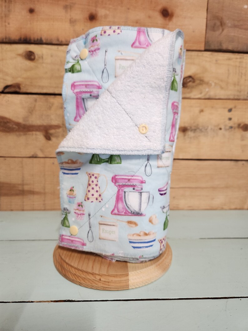 Pu&ograve; includere: Un porta asciugamani da cucina in tessuto blu e bianco con un mixer rosa e argento, una caraffa e cupcake stampati su di esso. Il porta asciugamani &egrave; in tessuto e ha una fodera in spugna bianca. Il porta asciugamani &egrave; appoggiato su una base in legno.