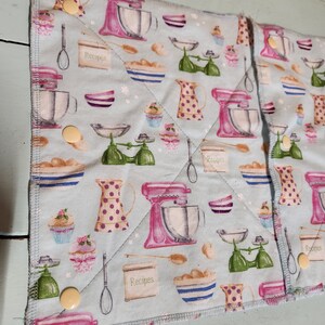 Pu&ograve; includere: Due presine in tessuto blu e bianco a tema cucina. Il tessuto presenta un motivo ripetitivo di planetarie rosa, bilance verdi e ciotole con la scritta "Ricette" stampata su di esse.