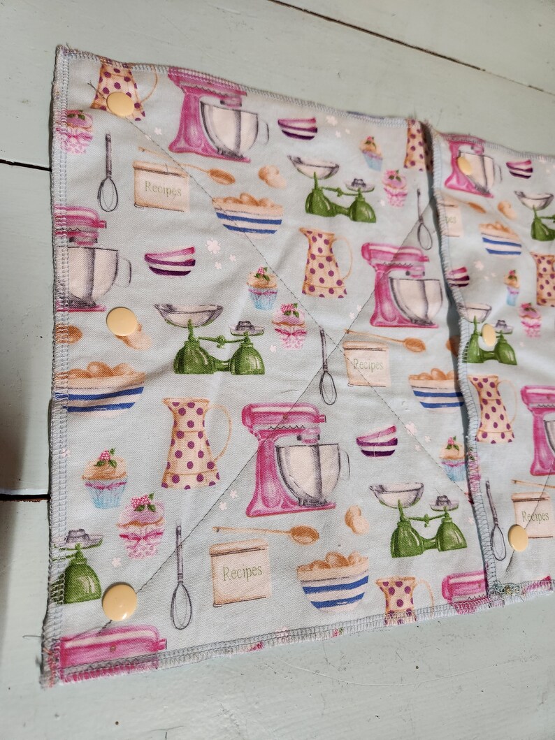 Pu&ograve; includere: Due presine in tessuto blu e bianco a tema cucina. Il tessuto presenta un motivo ripetitivo di planetarie rosa, bilance verdi e ciotole con la scritta "Ricette" stampata su di esse.