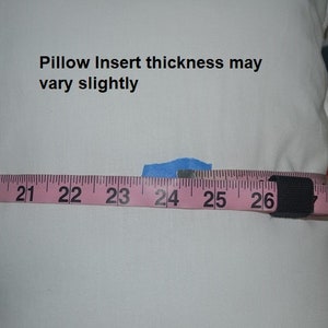 May include: A white pillow with a pink measuring tape wrapped around it. The tape reads "20, 21, 22, 23, 24, 25, 26". The text "Pillow Insert thickness may vary slightly" is printed on the pillow.