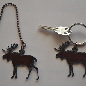 May include: Two moose silhouette keychains, one on a brown chain and the other on a silver key ring. The keychain on the ring is attached to a silver key.