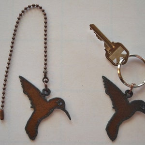 May include: Two hummingbird-shaped metal ornaments. One is attached to a pull chain, the other to a key ring with a key. The hummingbirds are brown with detailed wing and beak designs. The key is silver and gold.
