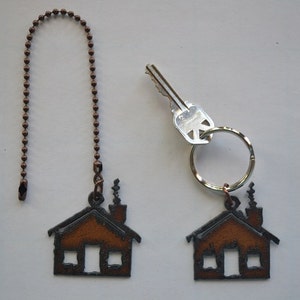May include: Two keychains with a house-shaped charm. The charm is made of metal and has a brown patina. One keychain has a copper chain and the other has a silver key ring.