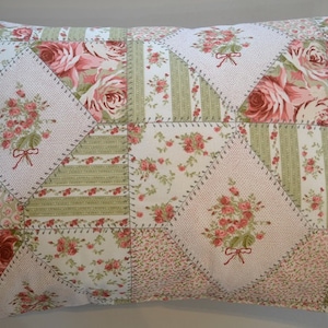 May include: A rectangular pillow with a patchwork design. The pillow features a variety of floral patterns in shades of pink, green, and white. The patchwork includes large roses, floral bouquets, and striped sections. The pillow is approximately 20 inches long and 12 inches wide.