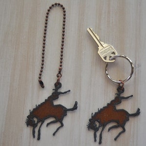 May include: Two rustic brown metal cowboy riding a bucking horse charms. One charm is attached to a copper chain and the other is attached to a key ring.
