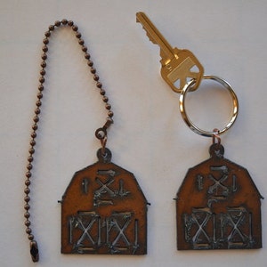 May include: Two rustic brown metal barn-shaped keychains. One keychain has a silver chain and the other has a silver key ring. Both keychains have a white painted design of a barn with a roof and a door.