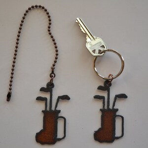 May include: Two golf bag keychains, one on a brown chain and the other on a silver key ring. Both keychains are made of metal and have a brown painted finish. The golf bag is outlined in black and has golf clubs sticking out of the top.