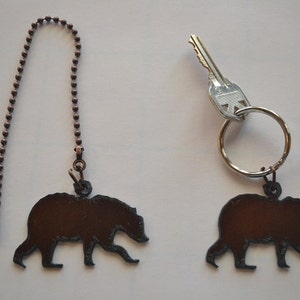 May include: Two brown metal bear silhouette keychains. One keychain is attached to a silver key ring with a key. The other keychain is attached to a silver chain.