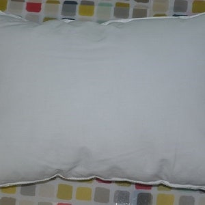 May include: A white pillow with a white border. The pillow is lying flat on a colorful patterned surface.