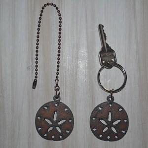 May include: Two decorative keychains and a ceiling fan pull chain. Each features a bronze-colored sand dollar charm. One keychain holds a key. The pull chain has a ball chain. The background is a light wood grain.