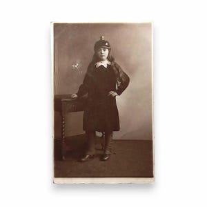 May include: A vintage sepia-toned photograph featuring a person with long hair wearing a dark coat, a light-coloured ruffled collar, and a rounded cap. The person stands with one hand on their hip next to a small wooden table with a twisted leg.