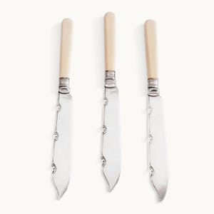 May include: Three silver-plated knives featuring smooth, cream-colored cylindrical handles and ornate, decorative scrollwork detailing along the edge of the metal blades. Each knife includes a polished silver ferrule connecting the handle to the blade.