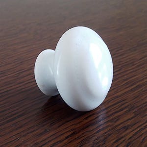 Op de afbeelding: Twee witte, ovaalvormige kastknoppen. Eén knop is kleiner en bevestigd aan een grotere knop. De knoppen hebben een gladde, glanzende afwerking en staan op een donkerbruin houten oppervlak.