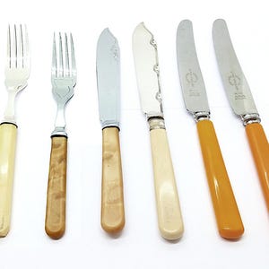May include: A set of six antique silverware pieces, including two forks and four knives. The forks have silver tines and cream-colored handles. The knives have silver blades and handles in cream and amber colors. The silverware is arranged on a white surface.