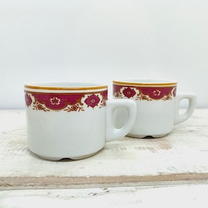 May include: Two white ceramic mugs featuring a decorative band around the upper rim. The band consists of a vibrant magenta floral pattern outlined in gold, set against a white background. Each mug has a sturdy handle and a smooth, glossy finish.