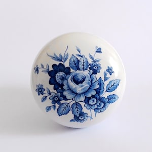 May include: A white ceramic cabinet knob with a blue floral pattern. The design shows a cluster of blue flowers and leaves. The knob is round and has a glossy finish. Perfect for adding a touch of elegance to furniture.