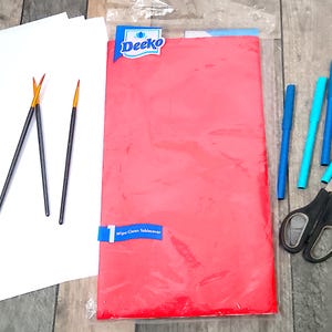 Op de afbeelding: Een rechthoekig rood Deeko tafelkleed in een doorzichtige plastic verpakking, vergezeld van drie dunne penselen met zwarte stelen, drie blauwe viltstiften en een zwarte schaar met grijze handgrepen.