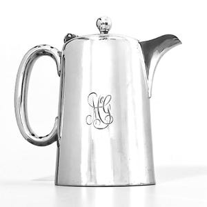 Op de afbeelding: Een zilverkleurige koffiepot met een gebogen handvat en een tuit. De pot heeft een deksel met een kleine knop en een monogram op de voorkant. De pot is gemaakt van een glanzend, reflecterend metaal.