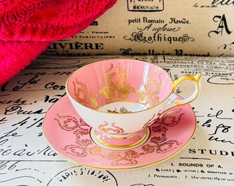May include: A pink and gold teacup and saucer set with a delicate floral pattern. The teacup has a white interior and a gold rim. The saucer has a pink rim and a gold floral design.