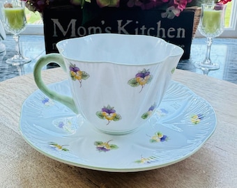 Peut inclure: Une tasse à thé et une soucoupe blanches avec un bord vert et un motif floral. La tasse à thé a un bord festonné et la soucoupe a un bord festonné. La tasse à thé et la soucoupe sont posées sur une table en bois. Le texte "Mom's Kitchen" est visible sur une boîte en bois à l'arrière-plan.