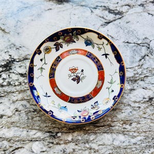 May include: A white saucer with a detailed floral design. The saucer has a blue and gold rim with a red and gold inner ring. The center of the saucer features a floral motif. The saucer is placed on a marble surface.