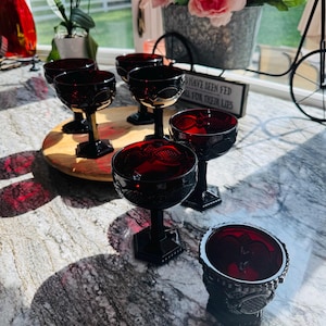 May include: A collection of six deep red glass goblets featuring a textured, pressed pattern on the bowls and hexagonal bases. The goblets are arranged on a round wooden board, with some positioned on the board and others resting on the surface.