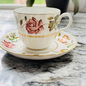 May include: A white porcelain teacup and saucer set featuring a delicate hand-painted pink rose motif with green leaves and gold trim accents along the rim, handle, and base. The teacup has a curved handle and a smooth, glossy finish throughout.