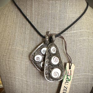 May include: A silver-toned pendant necklace featuring two pendants with a textured surface. Each pendant has white spiral shell accents. The necklace is displayed on a neutral-toned surface. A tag reads "Lou Zeldis."