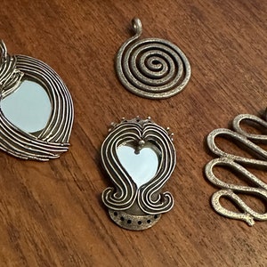 May include: Four silver-toned pendants with unique designs. One oval pendant has a mirror center, while another is a spiral. The third is heart-shaped with a mirror, and the last is a wavy design. All are on a wooden surface.