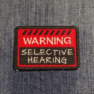 May include: A rectangular embroidered patch attached to blue denim fabric. The patch features a red top section with black diagonal stripes and the white text WARNING, and a black bottom section with the white text SELECTIVE HEARING.
