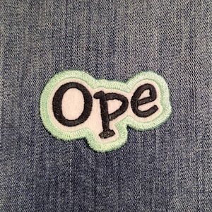 May include: An embroidered patch featuring the word Ope in black lettering with a white outline, set against a light mint green background. The patch is attached to a blue denim fabric surface with visible vertical seams and stitching details.