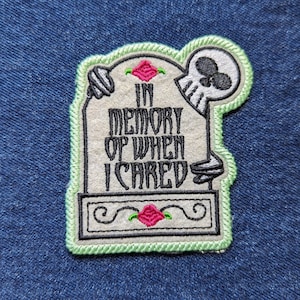 May include: An embroidered patch shaped like a tombstone with a light green border. It features a white skull, two pink roses with green leaves, and the black embroidered text "IN MEMORY OF WHEN I CARED" centered on a light gray background.